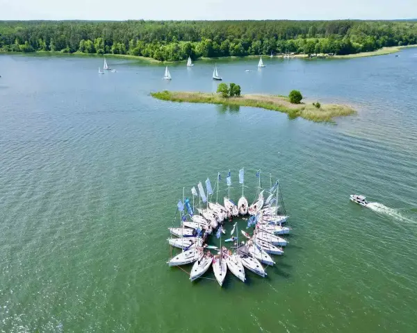 Żaglówki tworzą okrąg na spokojnym **lago**, otoczone zielonym lasem.