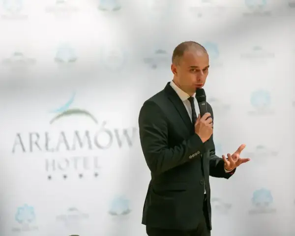 Mężczyzna w garniturze przemawia na tle logo Hotel Arłamów. Prawdopodobnie właściciel hotelu.