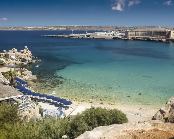 Rajska zatoka na Malcie z turkusową wodą, piaszczystą plażą i rzędem niebieskich parasoli. W oddali widać hotel i port.