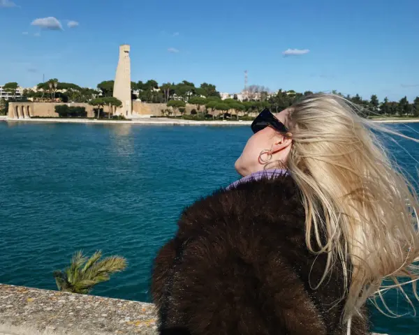 Kobieta z blond włosami podziwia pomnik w Brindisi. W tle błękitne morze i błękitne niebo.