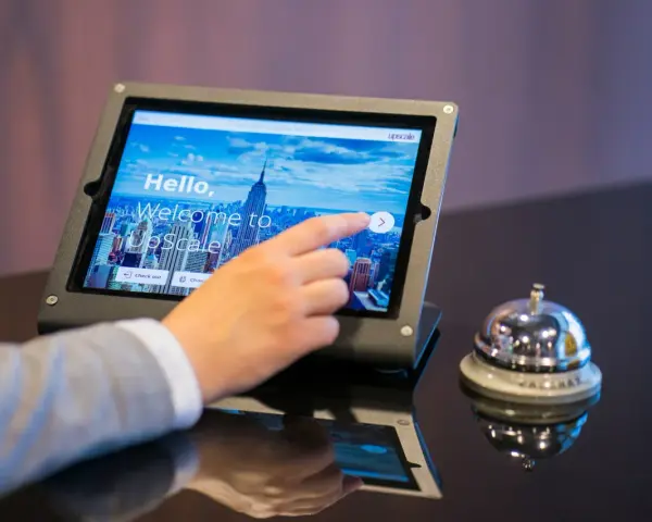 Dłoń dotyka ekranu tabletu, rozpoczynając proces zameldowania w hotelu. Obok stoi dzwonek hotelowy.