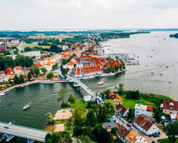 Malownicze miasta na Mazurach z widokiem na jezioro i liczne łodzie. Biały most łączy brzegi, a czerwone dachy domów tworzą urokliwy krajobraz.