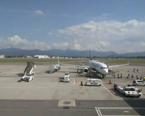Samolot Ryanair na płycie lotniska Bergamo. W tle góry, na pierwszym planie schody, pojazdy obsługi naziemnej i pasażerowie.