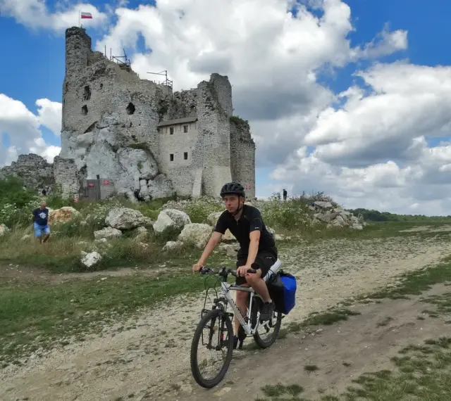 Młody rowerzysta przemierza malownicze **Jura Krakowsko-Częstochowska szlaki**, mijając ruiny zamku na tle błękitnego, chmurnego nieba.
