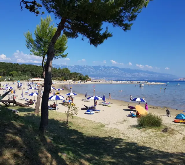 Słoneczny dzień na plaży, pełnej parasoli i ludzi. Woda jest spokojna, a w oddali widać góry. To prawdziwy raj na ziemi.
