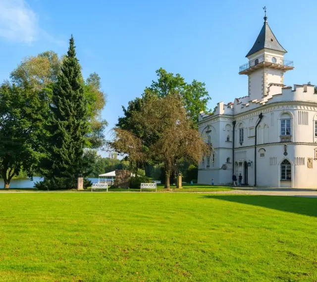 Biały pałac z wieżą, otoczony zielenią, nad jeziorem. Jeden z urokliwych zamków na Mazowszu.