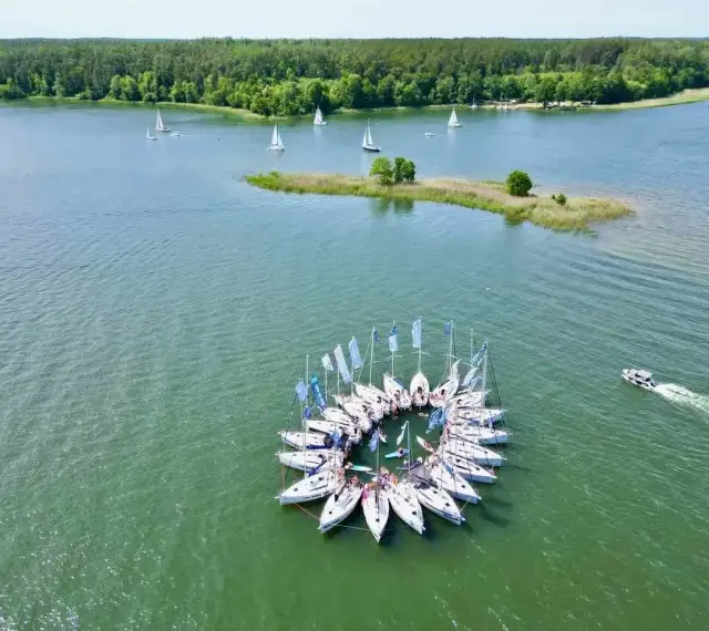 Żaglówki tworzą okrąg na spokojnym **lago**, otoczone zielonym lasem.