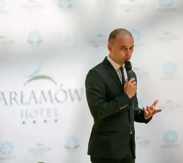 Mężczyzna w garniturze przemawia na tle logo Hotel Arłamów. Prawdopodobnie właściciel hotelu.