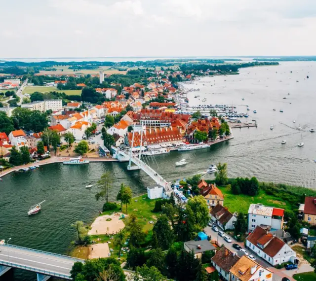 Malownicze miasta na Mazurach z widokiem na jezioro i liczne łodzie. Biały most łączy brzegi, a czerwone dachy domów tworzą urokliwy krajobraz.