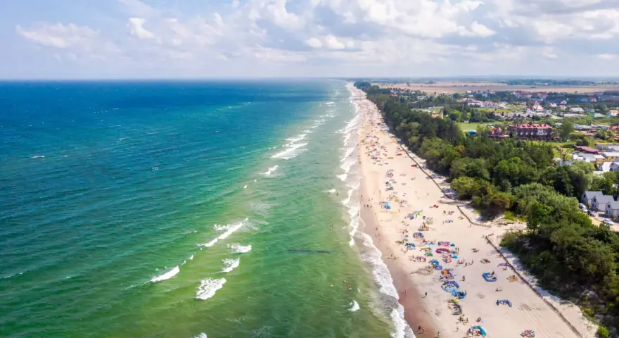 Słoneczny dzień na plaży w Sarbinowie nad morzem. Tłumy turystów odpoczywają na piasku, kąpią się w morzu i spacerują wzdłuż brzegu.