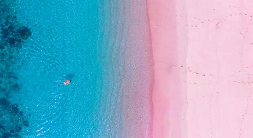 Widok z lotu ptaka na turkusowe morze i różową plażę w Grecji. Ktoś pływa w krystalicznie czystej wodzie.