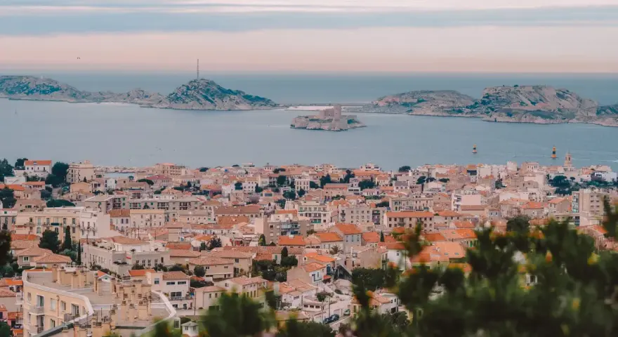 Widok na miasto z dachami i morzem w tle, z widocznym zamkiem na wyspie. To piękne **Marseille**.