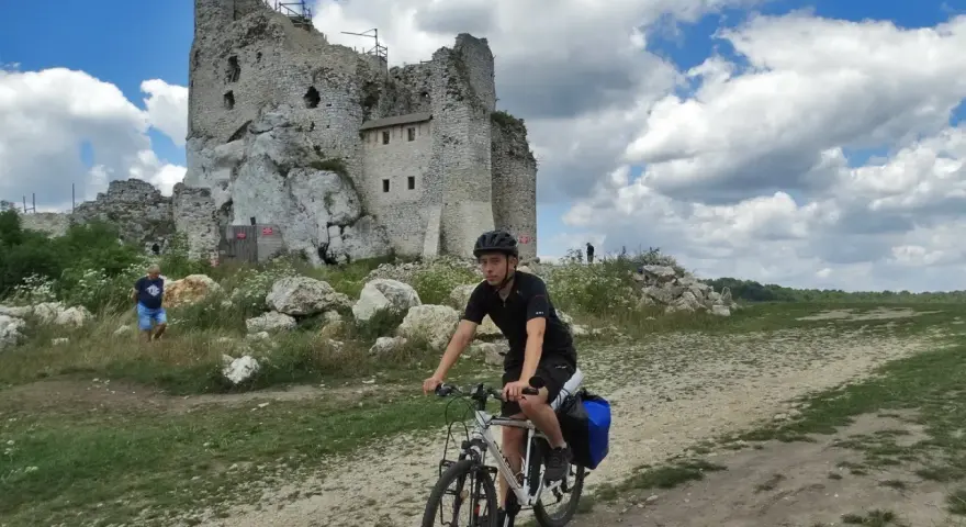 Młody rowerzysta przemierza malownicze **Jura Krakowsko-Częstochowska szlaki**, mijając ruiny zamku na tle błękitnego, chmurnego nieba.