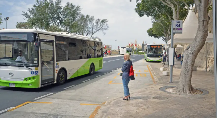 Kobieta czeka na autobus na Malcie. W tle widać autobusy, w tym jeden jadący w kierunku lotniska.