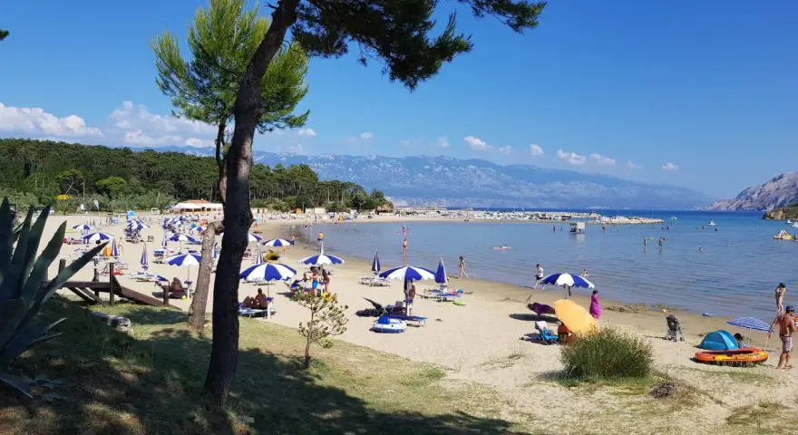 Słoneczny dzień na plaży, pełnej parasoli i ludzi. Woda jest spokojna, a w oddali widać góry. To prawdziwy raj na ziemi.