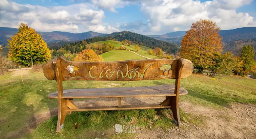 Drewniana ławka z napisem "Ciechocinek" na tle jesiennych gór i zielonych wzgórz. Widok na Beskid Sądecki.
