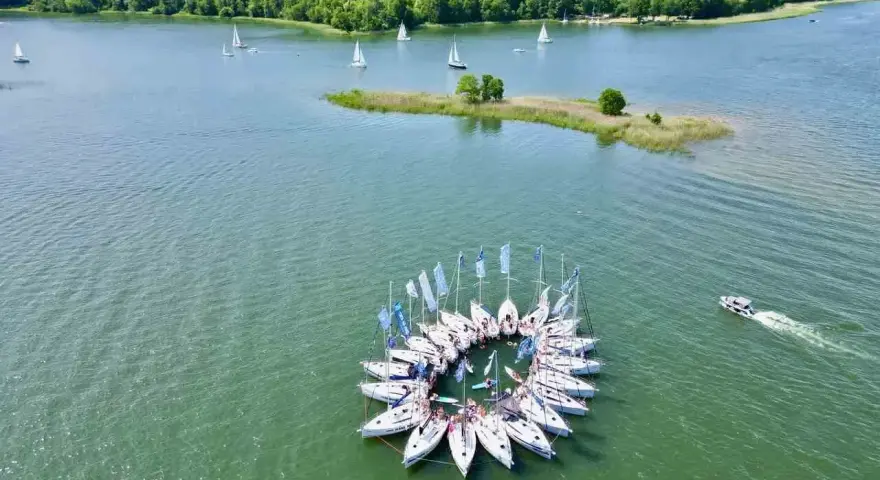 Żaglówki tworzą okrąg na spokojnym **lago**, otoczone zielonym lasem.