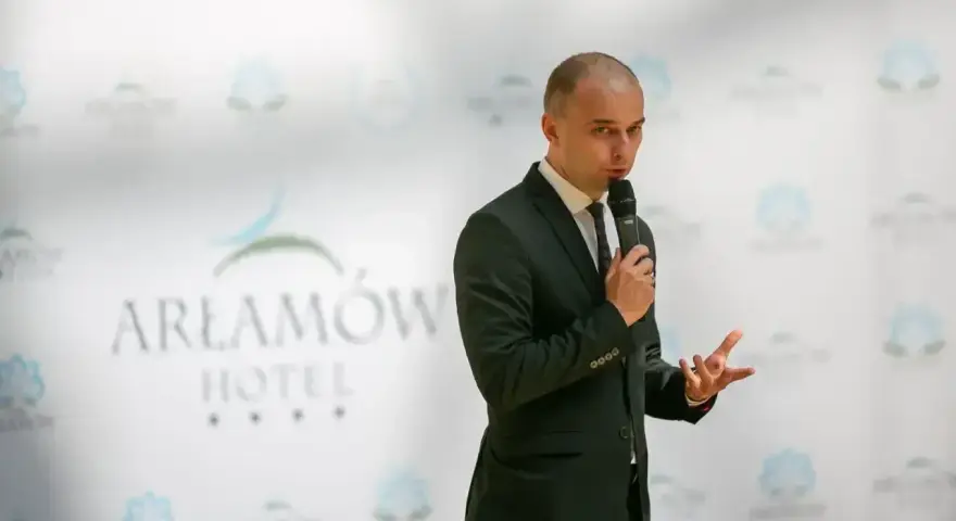 Mężczyzna w garniturze przemawia na tle logo Hotel Arłamów. Prawdopodobnie właściciel hotelu.