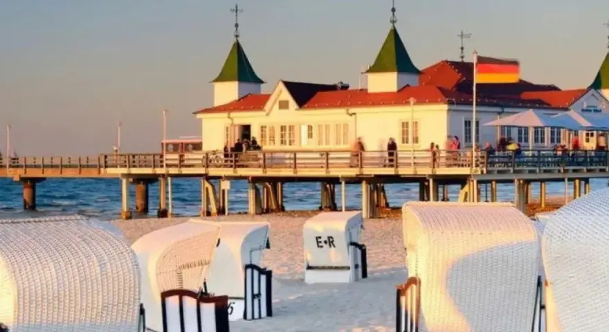 Plaża z koszami i molo, gdzie widać budynek restauracji i niemiecką flagę. Atrakcje po niemieckiej stronie Świnoujścia zachęcają do spacerów.