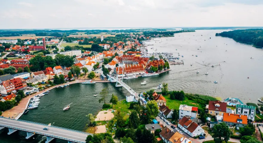 Malownicze miasta na Mazurach z widokiem na jezioro i liczne łodzie. Biały most łączy brzegi, a czerwone dachy domów tworzą urokliwy krajobraz.