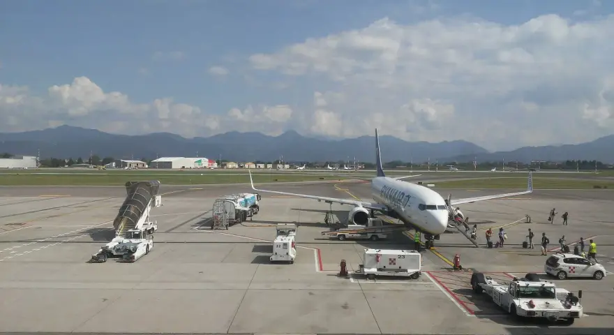 Samolot Ryanair na płycie lotniska Bergamo. W tle góry, na pierwszym planie schody, pojazdy obsługi naziemnej i pasażerowie.