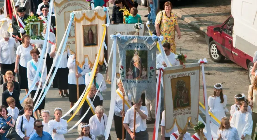 Procesja z banerami religijnymi, w tym z napisem "Boże Ciało 26". Ludzie niosą obrazy i kwiaty.