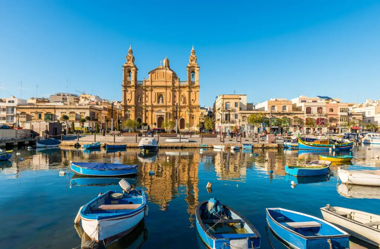 Malta: kolorowe łodzie kołyszą się w porcie przed barokową bazyliką. Idealne miejsce na wakacje, nie lotniska.