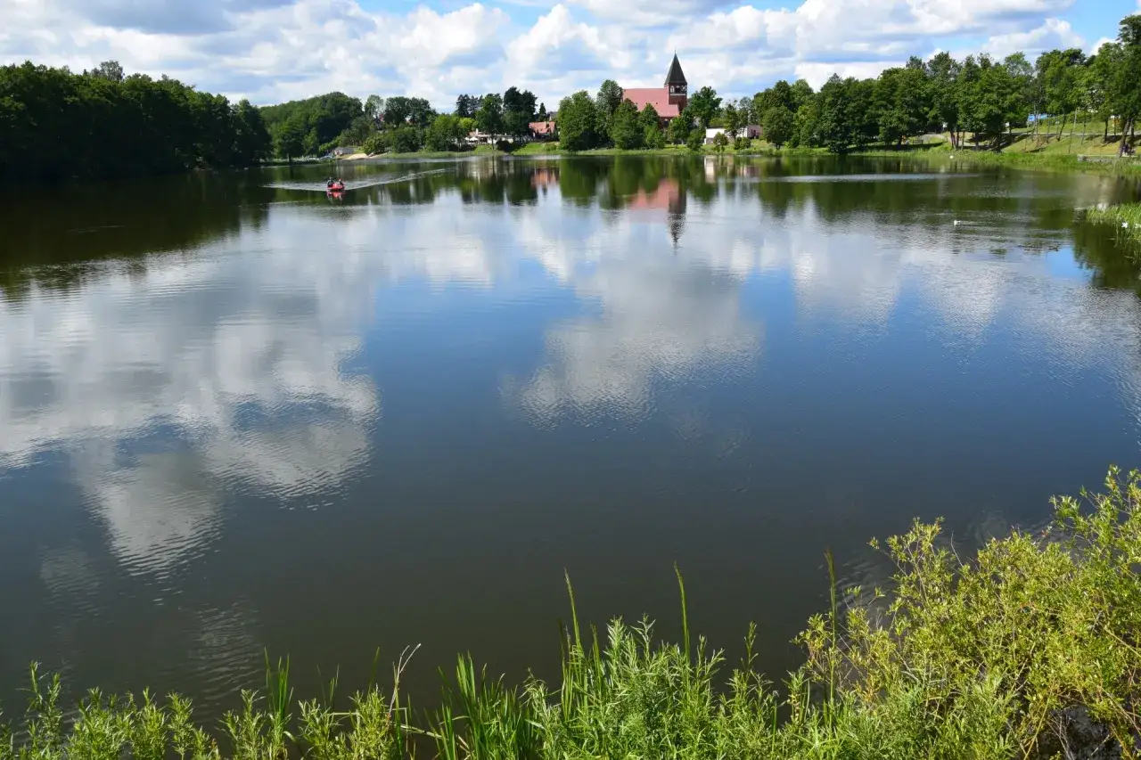 Spokojne jeziora na Kaszubach, odbijające chmury. W oddali widać kościół i zielone drzewa.