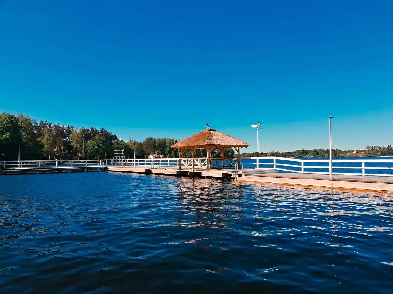 Drewniana plaża z altaną i białymi barierkami nad spokojnym jeziorem. Idealne miejsce na relaks w narie resort & spa.
