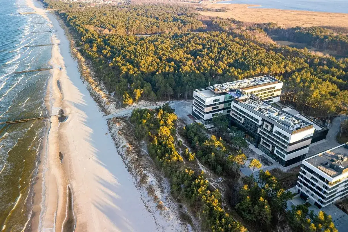 Nowoczesny hotel 5 gwiazdkowy nad morzem, otoczony lasem sosnowym, z widokiem na plażę i fale.