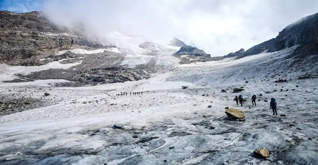 Grupa turystów wspina się po lodowcu w kierunku majestatycznego Monte Rosa. Chmury otulają szczyty, tworząc mistyczną atmosferę.