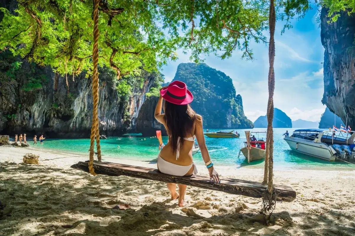 Kobieta w białym bikini i różowym kapeluszu siedzi na huśtawce na plaży. W tle turkusowa woda, łodzie i wapienne klify. Idealne miejsce na wakacje w Tajlandii w sierpniu.