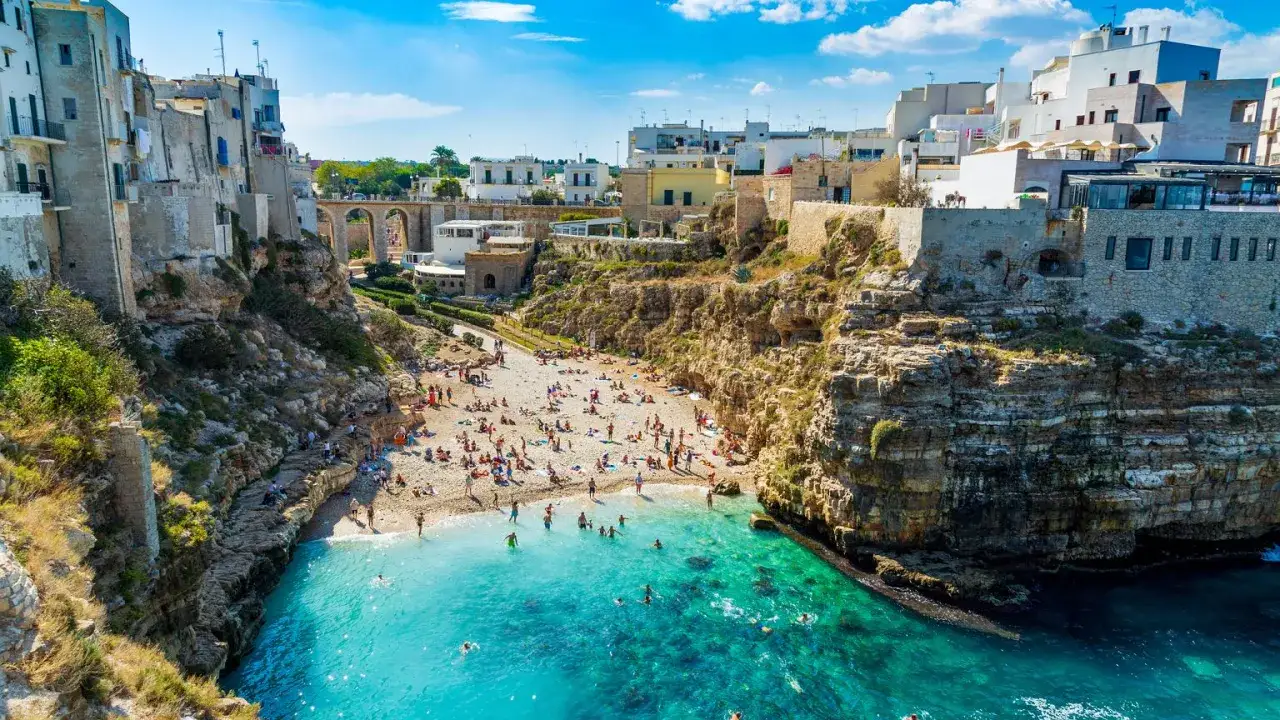 Malownicze bari plaże z białymi budynkami na klifach i turkusową wodą. Ludzie odpoczywają na kamienistej plaży i kąpią się w morzu.