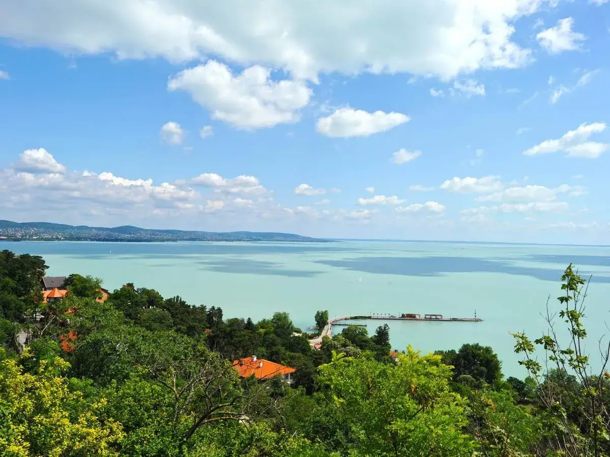 Widok na turkusowe jezioro Balaton z zielonymi wzgórzami i domami. Na wodzie widać molo i żaglówki.