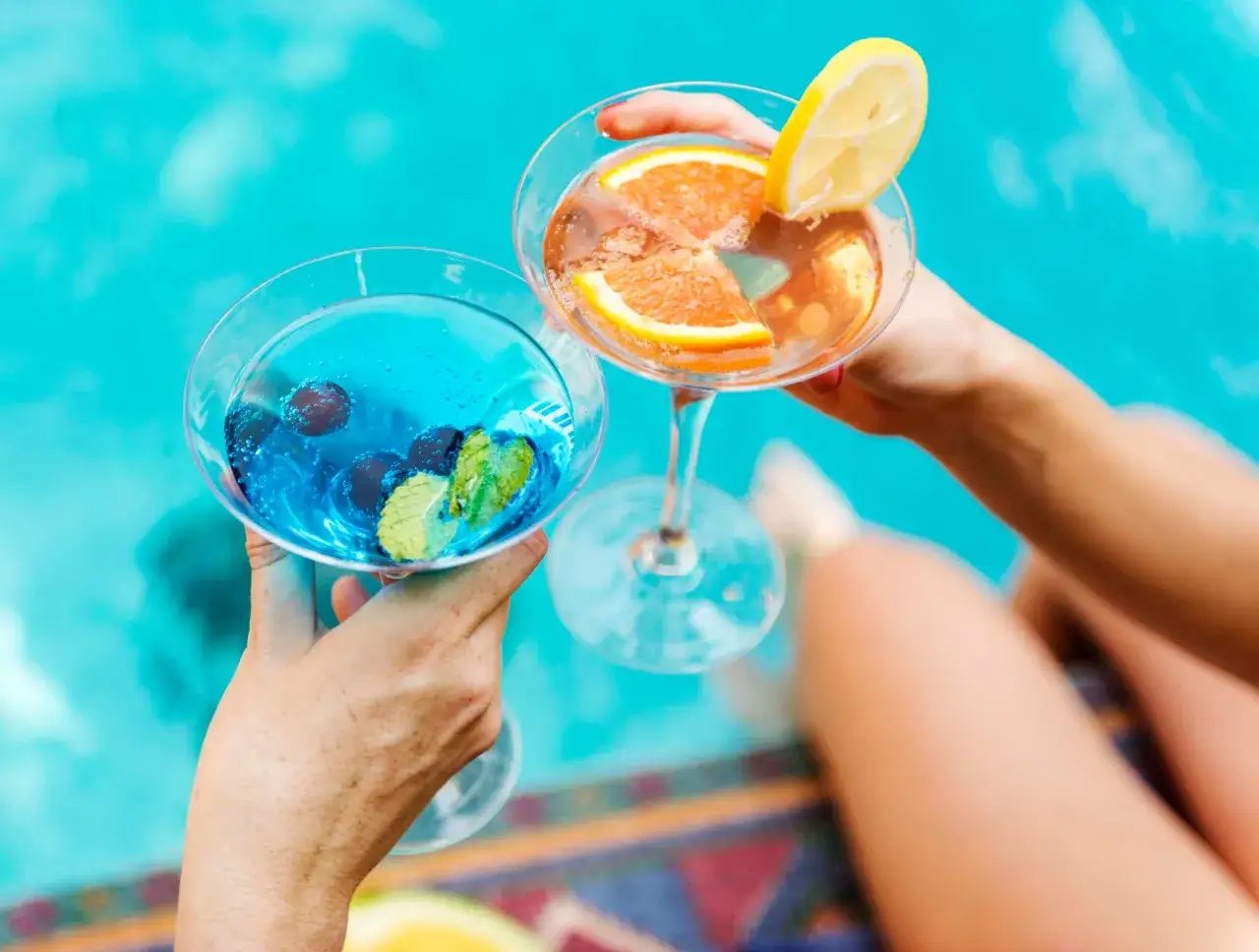 Dwie ręce unoszą kieliszki z kolorowymi drinkami przy basenie. Idealny all inclusive soft na wakacjach.