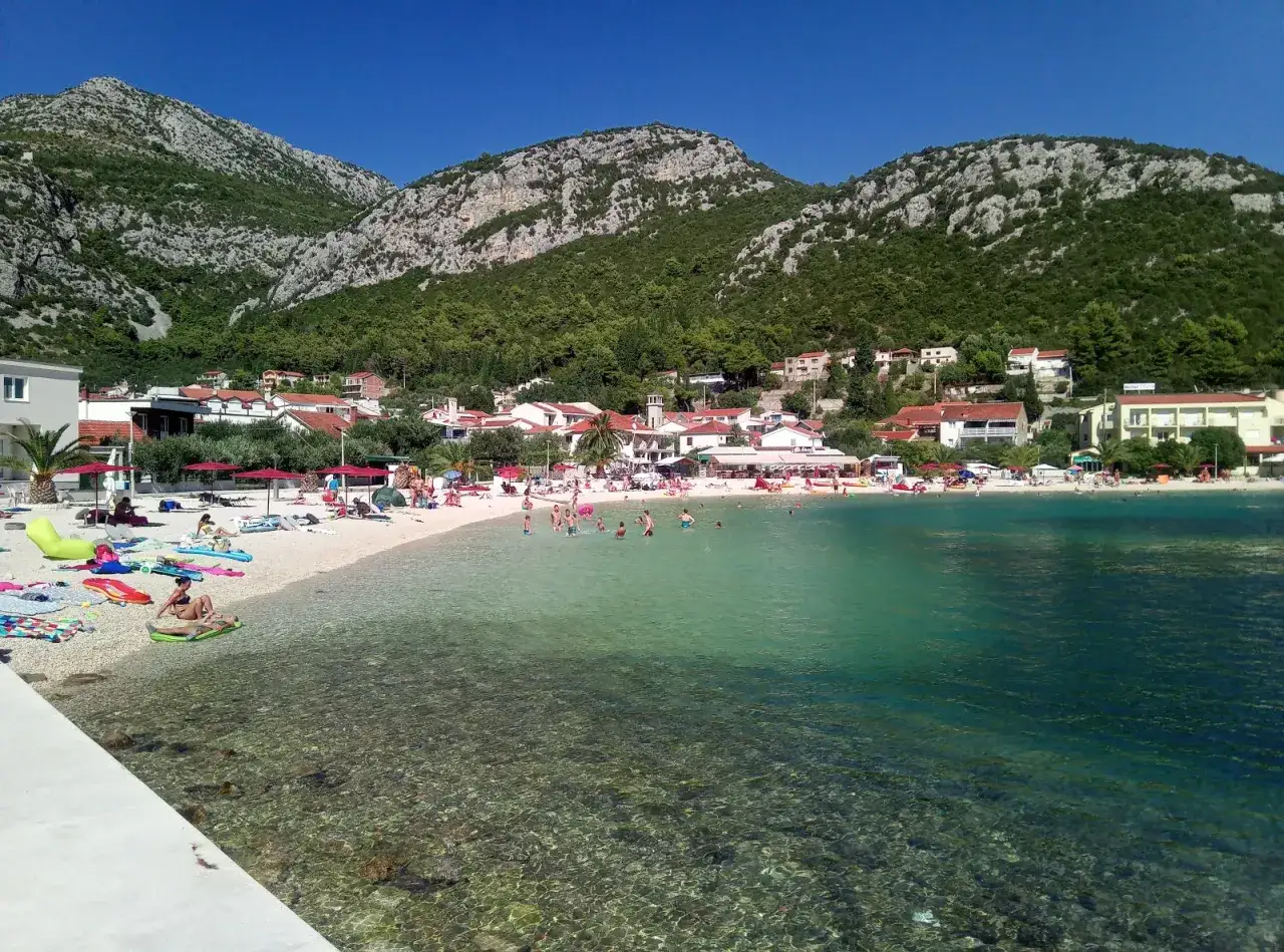 Słoneczna plaża z turkusową wodą, góry w tle. Ludzie odpoczywają na piasku i kąpią się. W oddali widać miasteczko z białymi domami. Idealne miejsce na wakacyjny klek.