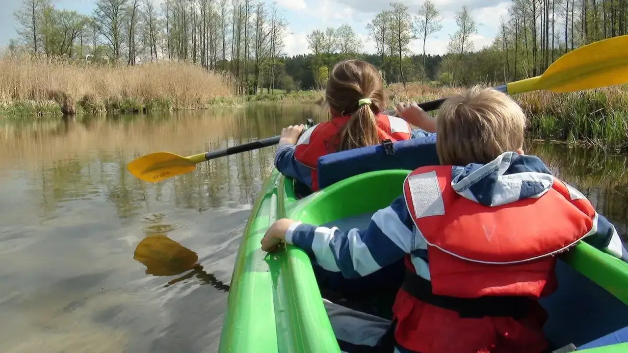 Dzieci w kamizelkach ratunkowych płyną kajakiem po spokojnym lago. Wiosła odbijają się w wodzie.