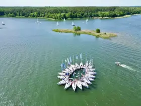 Żaglówki tworzą okrąg na spokojnym **lago**, otoczone zielonym lasem.