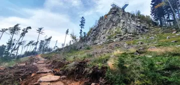 Kamienista ścieżka prowadzi ku skalistemu szczytowi z platformą widokową. W oddali widać las, a krajobraz przypomina trojak Lądek Zdrój.