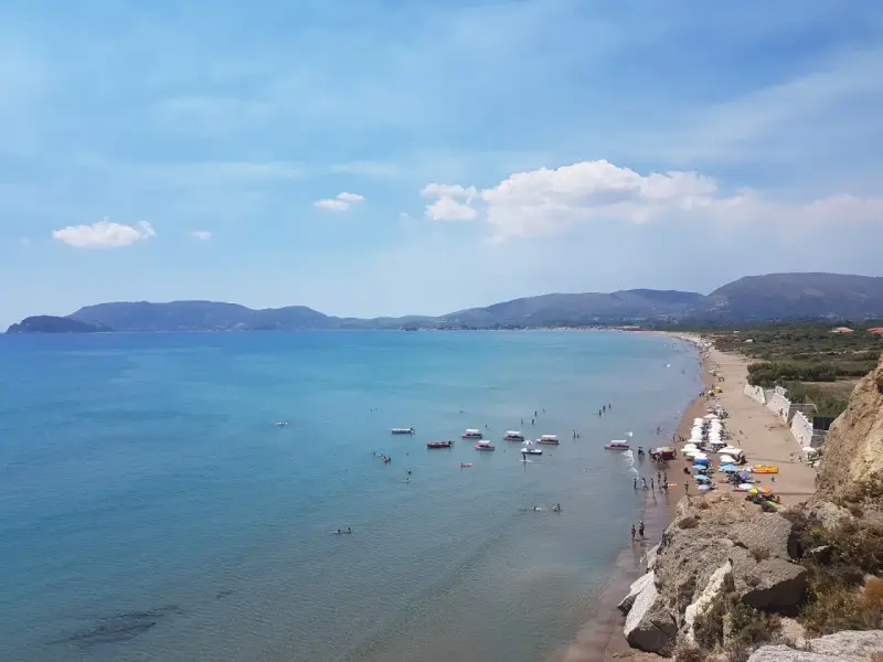 Słoneczny dzień na plaży Kalamaki. Ludzie pływają, opalają się i relaksują na piasku. Woda jest spokojna i turkusowa.