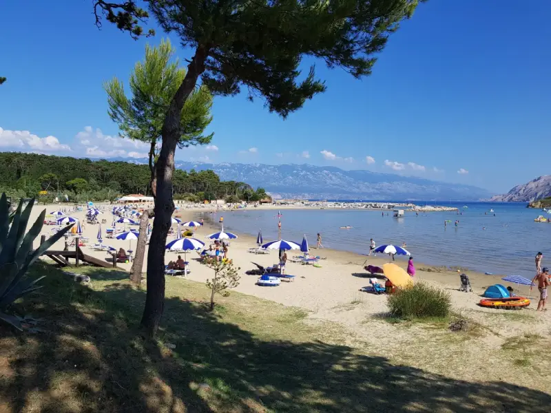 Słoneczny dzień na plaży, pełnej parasoli i ludzi. Woda jest spokojna, a w oddali widać góry. To prawdziwy raj na ziemi.