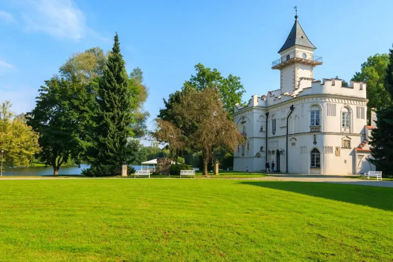 Biały pałac z wieżą, otoczony zielenią, nad jeziorem. Jeden z urokliwych zamków na Mazowszu.