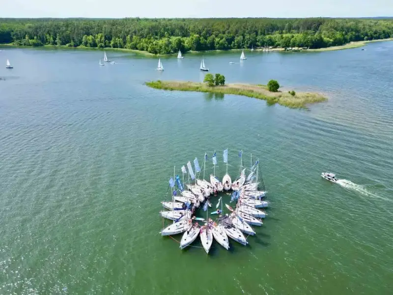 Żaglówki tworzą okrąg na spokojnym **lago**, otoczone zielonym lasem.