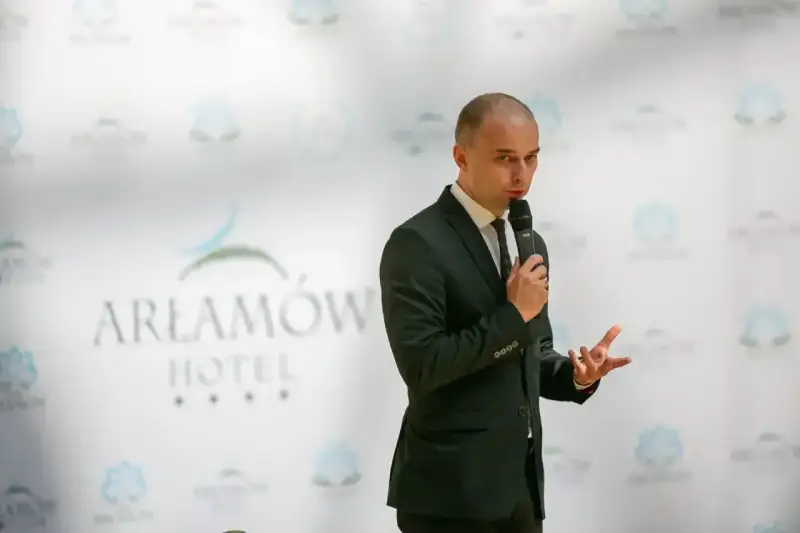 Mężczyzna w garniturze przemawia na tle logo Hotel Arłamów. Prawdopodobnie właściciel hotelu.