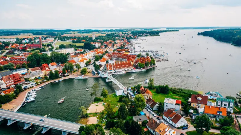 Malownicze miasta na Mazurach z widokiem na jezioro i liczne łodzie. Biały most łączy brzegi, a czerwone dachy domów tworzą urokliwy krajobraz.